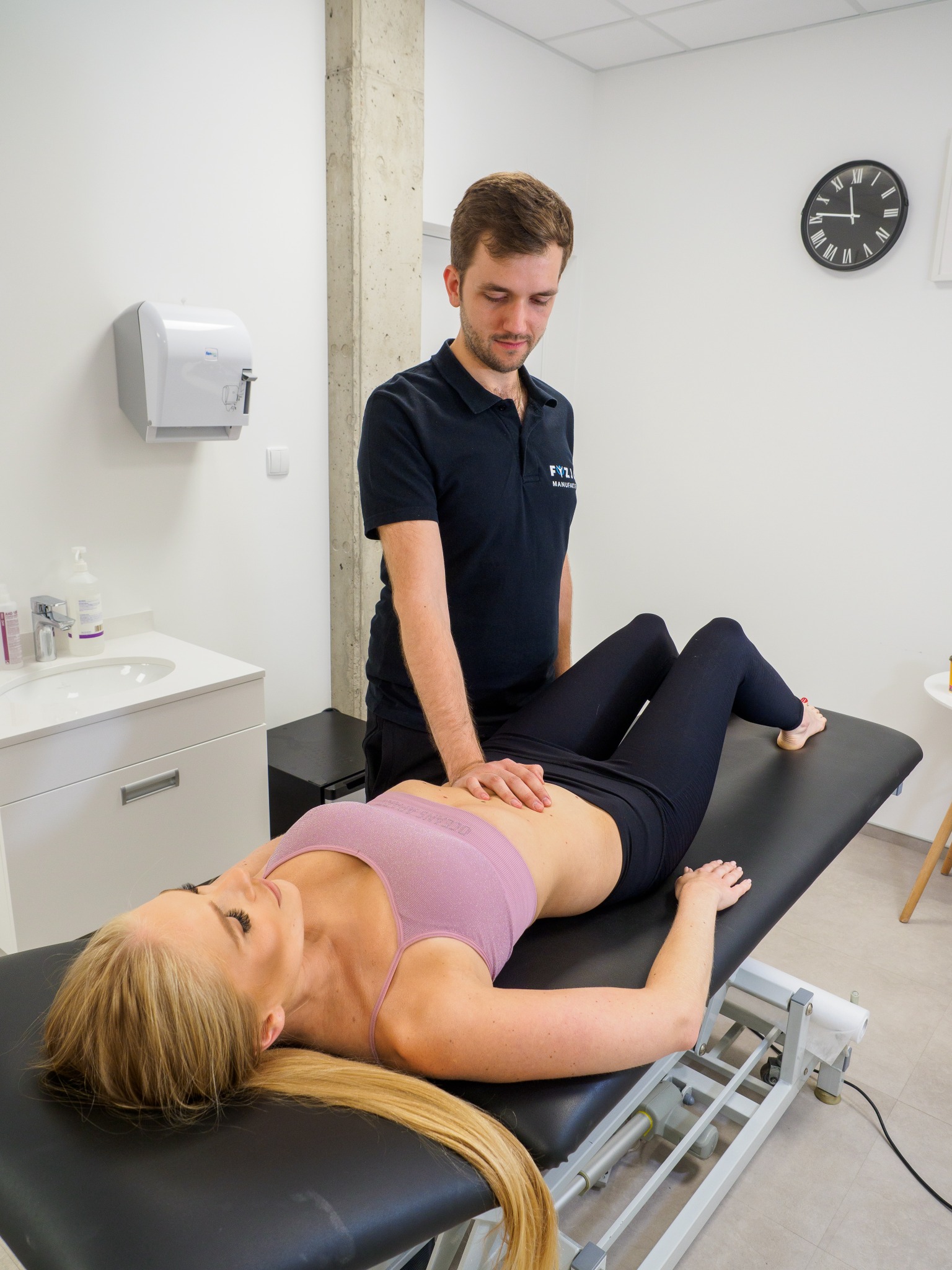 Kobieta leży na kozetce, terapeuta uciska jej brzuch dłonią, widoczny zegar na ścianie w tle.