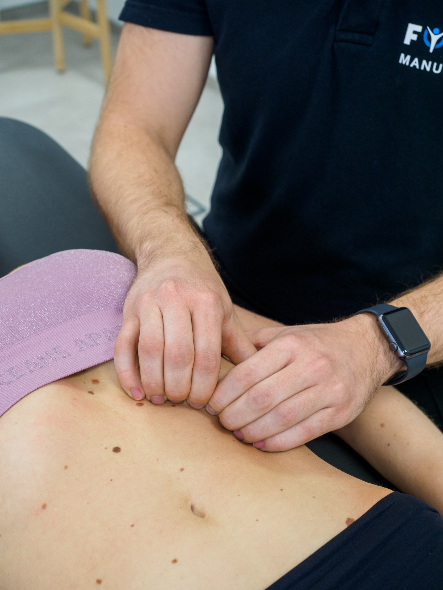 Pacjentka leżąca na stole do masażu, terapeuta uciska palcami okolice brzucha, widoczny zegarek na nadgarstku terapeuty i logo na koszulce.