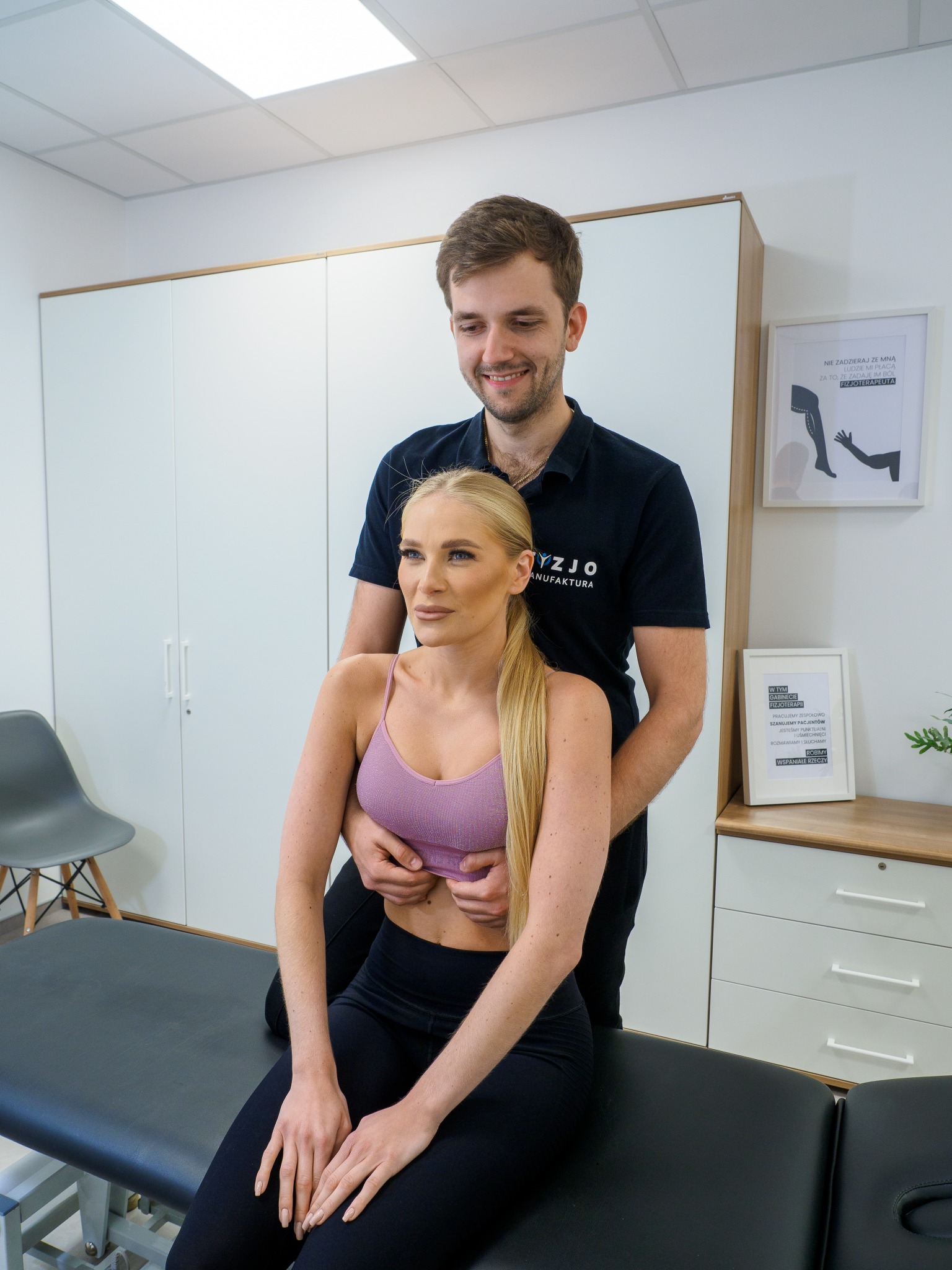Młody fizjoterapeuta w czarnym polo z logo firmy, delikatnie uciska klatkę piersiową pacjentki siedzącej na czarnym stole do masażu, w tle białe szafki i plakaty w ramkach.