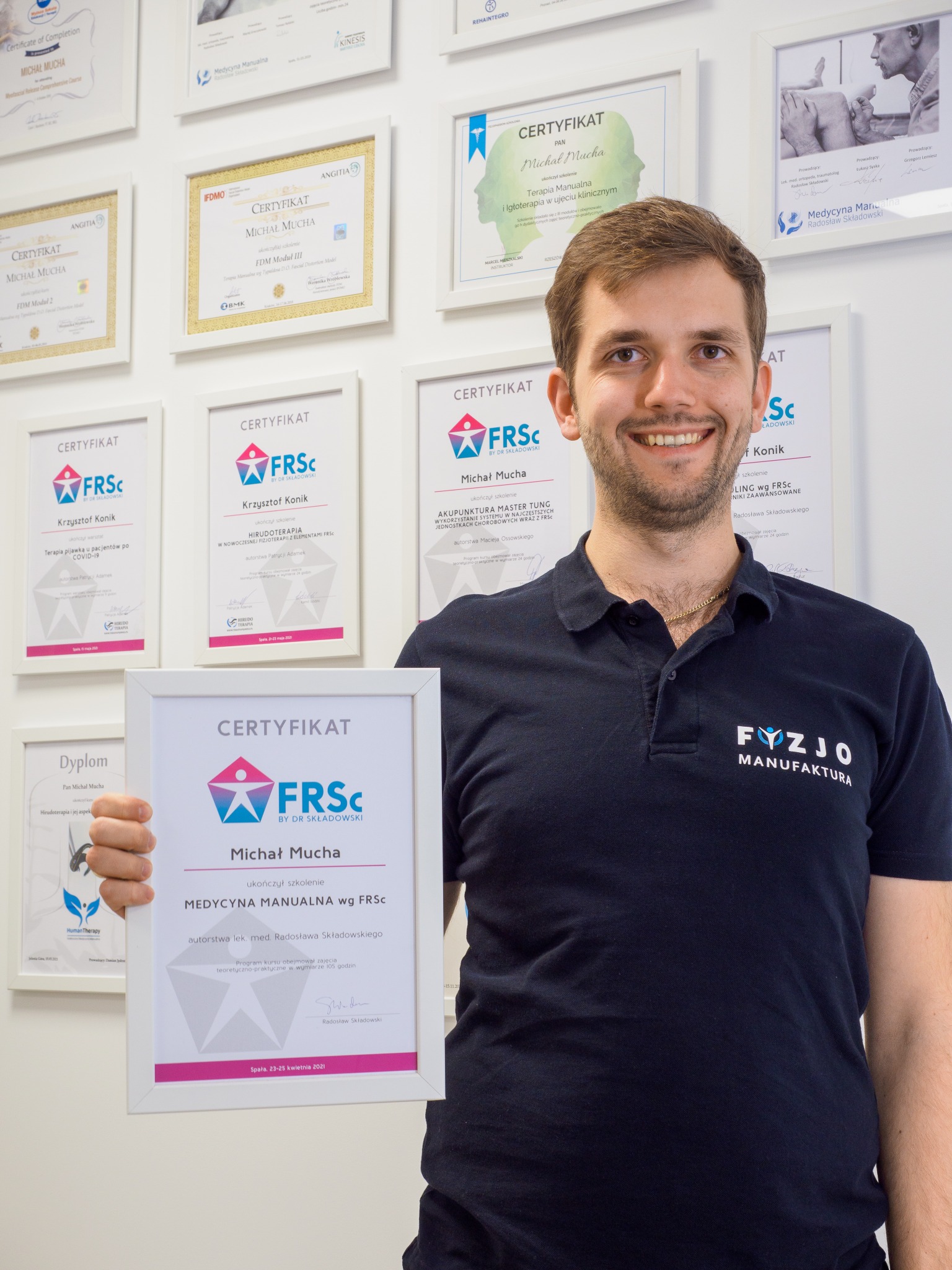 Uśmiechnięty fizjoterapeuta Michał Mucha prezentuje certyfikat ukończenia szkolenia z Medycyny Manualnej wg FRSc na tle ściany z licznymi dyplomami i certyfikatami, w tym z terapii manualnej...