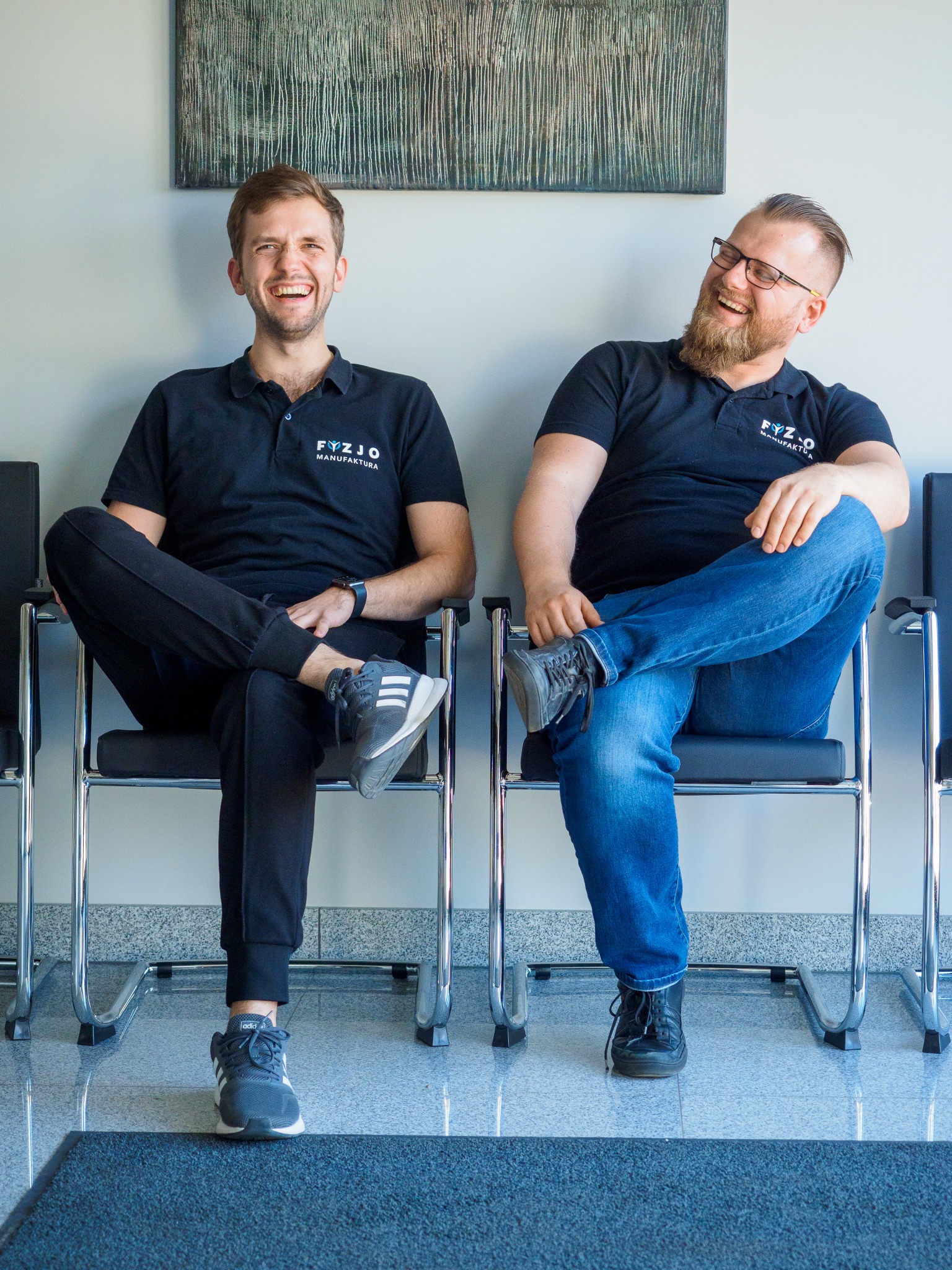 Dwóch uśmiechniętych mężczyzn w czarnych koszulkach z logo firmy siedzi na krzesłach w poczekalni, na ścianie wisi abstrakcyjny obraz.