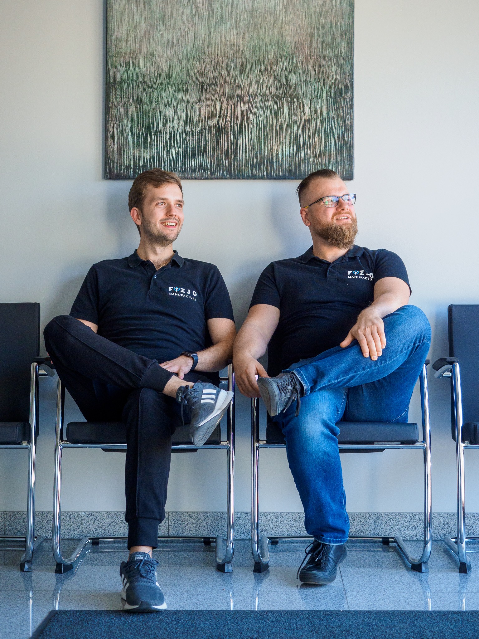 Dwóch uśmiechniętych mężczyzn w czarnych polówkach z logo firmy siedzi na krzesłach w poczekalni, z obrazem abstrakcyjnym na ścianie w tle.