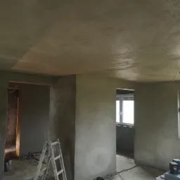 Wnętrze domu po nałożeniu świeżych tynków, widoczne ślady wilgoci na ścianach i suficie, drabina oparta o ścianę, okno z widokiem na zewnątrz, podłoga częściowo zabrudzona.