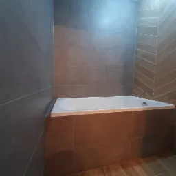 Wykończona łazienka z brązowymi i szarymi płytkami, z białą wanną w zabudowie i drewnopodobną podłogą.