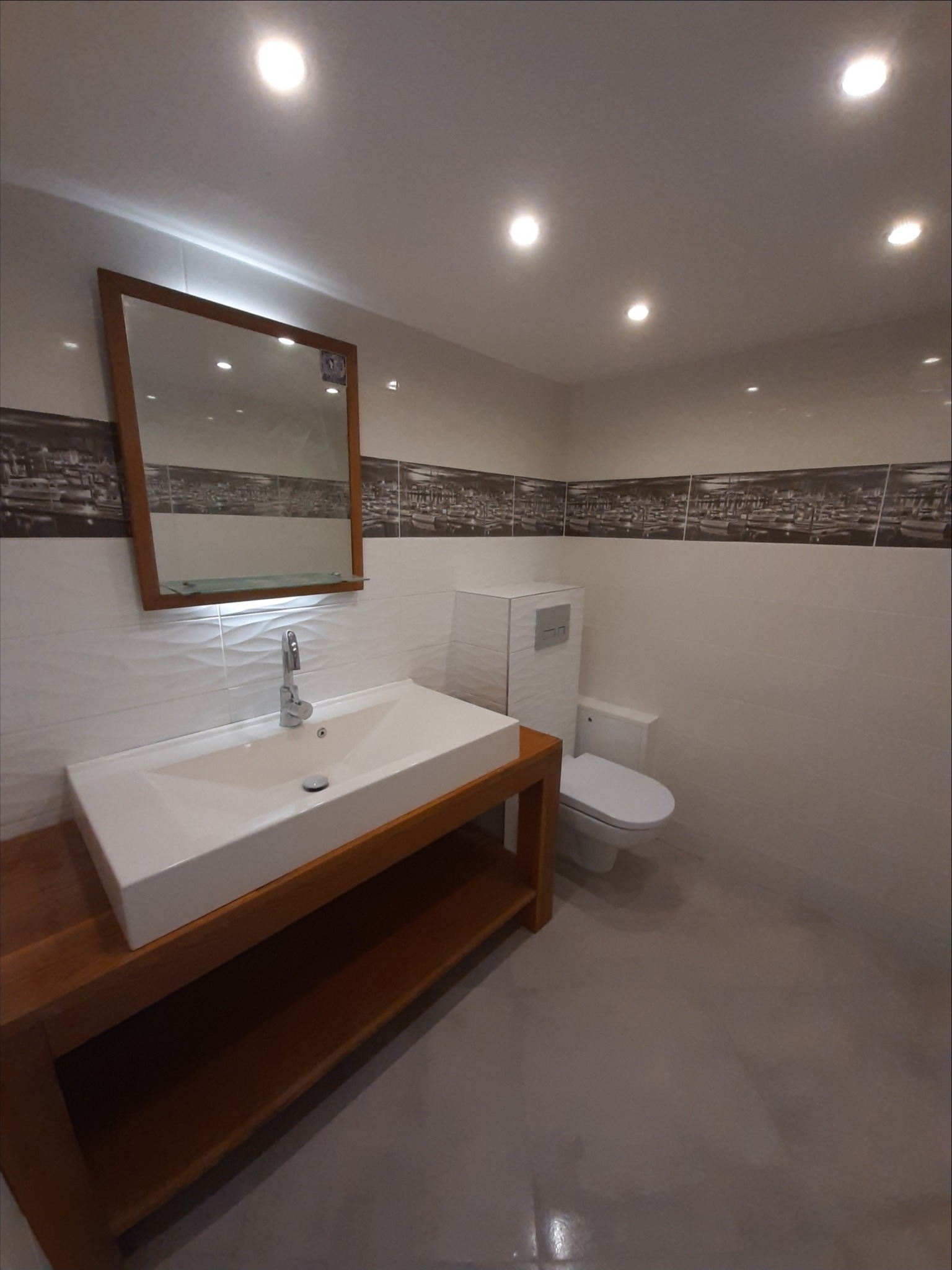 Nowoczesna łazienka z prostokątną umywalką nablatową na drewnianej szafce, lustro w drewnianej ramie, toaleta w zabudowie, płytki z motywem miasta nad umywalką i toaletą, oświetlenie punktowe.