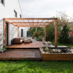 Drewniany taras z pergolą, stawem i meblami ogrodowymi przy białym domu, z elementami architektury krajobrazu w tle.