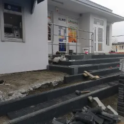 Montaż kostki brukowej przed wejściem do sklepu z bankomatem, widoczne narzędzia, poziomica, gumowy młotek i worki z materiałami budowlanymi.