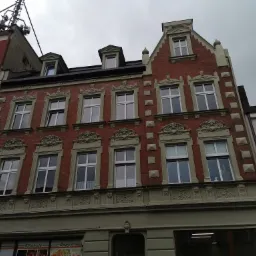 Fasada kamienicy z czerwonej cegły z ozdobnymi elementami architektonicznymi i oknami, widok z dołu na tle pochmurnego nieba.