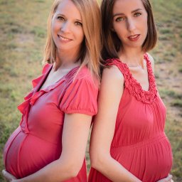 Monika Chocaj - Dwie uśmiechnięte kobiety w zaawansowanej ciąży, ubrane w czerwone sukienki, stoją plecami do siebie na tle rozmytej zieleni.