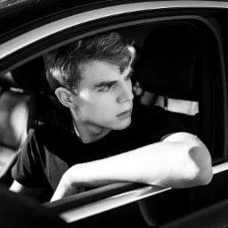 Czarno-białe zdjęcie młodego mężczyzny siedzącego w samochodzie, z ręką opartą o brzeg okna, spoglądającego w bok z zamyśloną miną.