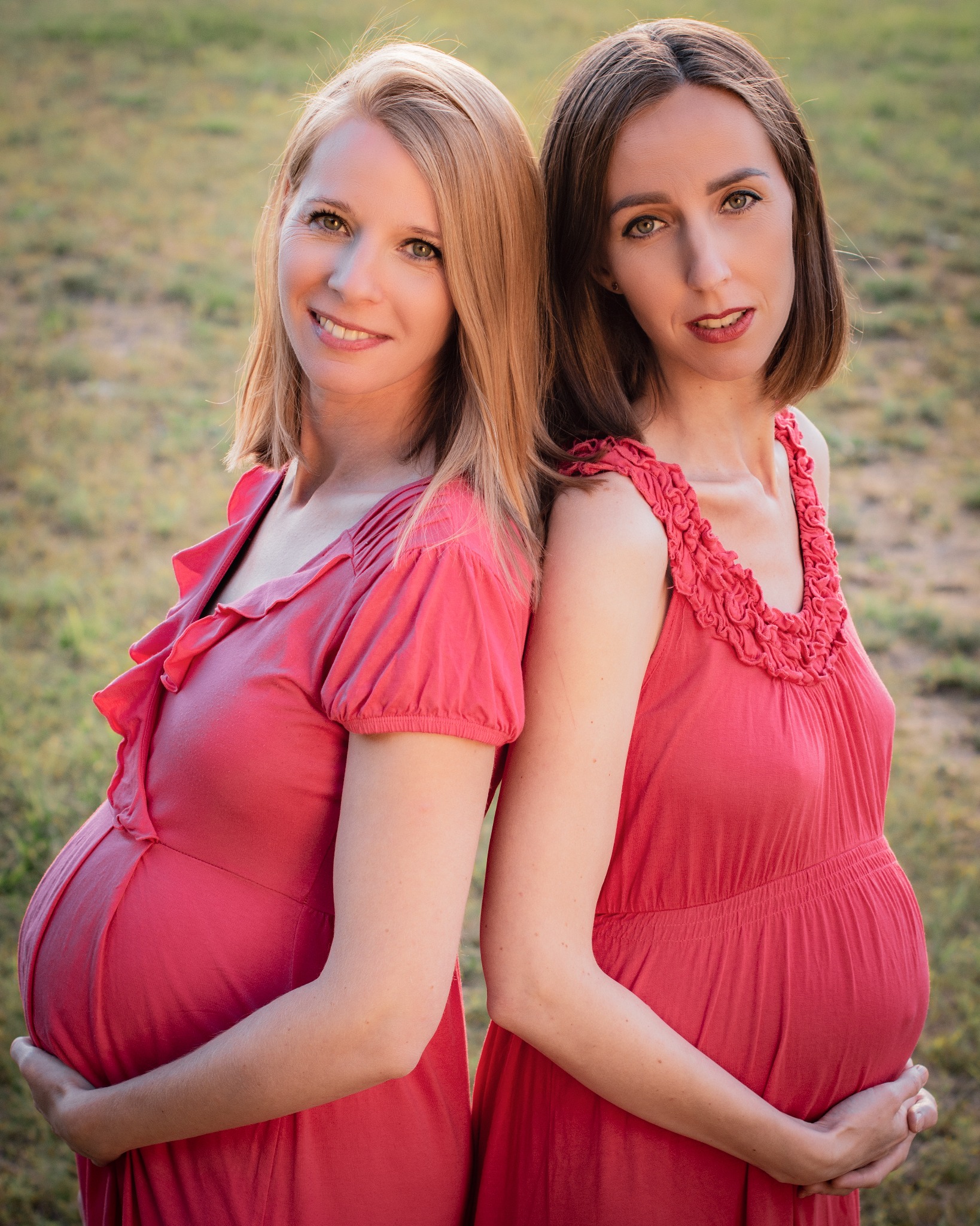 Dwie uśmiechnięte kobiety w zaawansowanej ciąży, ubrane w czerwone sukienki, stoją plecami do siebie na tle rozmytej zieleni.