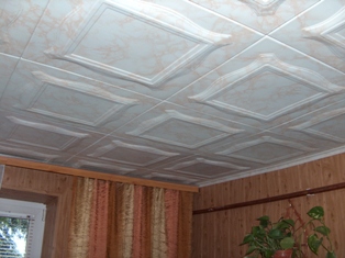 Kasetonowy sufit w odcieniach beżu i bieli, z wyraźnym wzorem kwadratów i dekoracyjnymi obramowaniami, widoczny fragment ściany z boazerią i zasłony w ciepłych barwach, roślina doniczkowa w rogu...