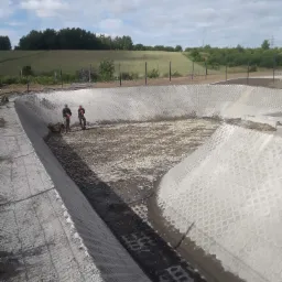 Budowa zbiornika retencyjnego wyłożonego ażurowymi płytami betonowymi, dwóch robotników z łopatami na dnie, widok z góry na tle zieleni.