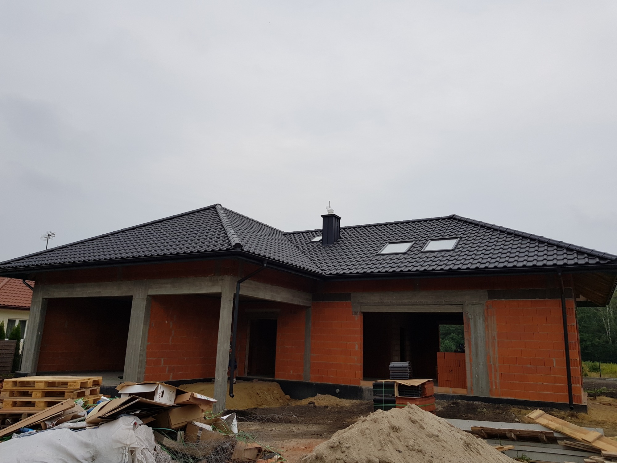Dom w stanie surowym otwartym w Opocznie z nowym, czarnym pokryciem dachowym i orynnowaniem. Widoczne okna dachowe i komin. Na pierwszym planie materiały budowlane i piasek.