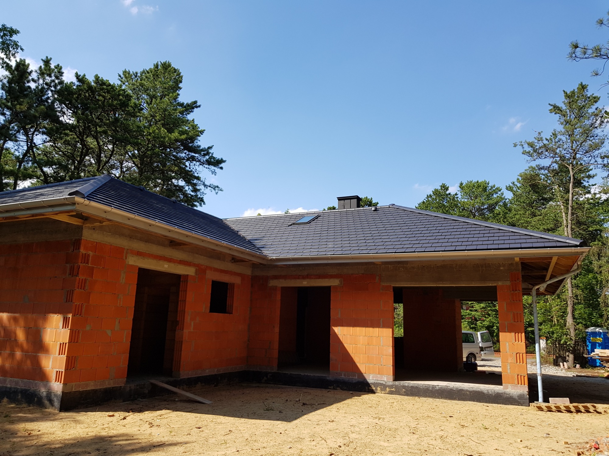 Niedokończony dom z cegły z nowym, ciemnoszarym dachem z dachówki ceramicznej, rynnami i oknem dachowym w świetle słonecznym.