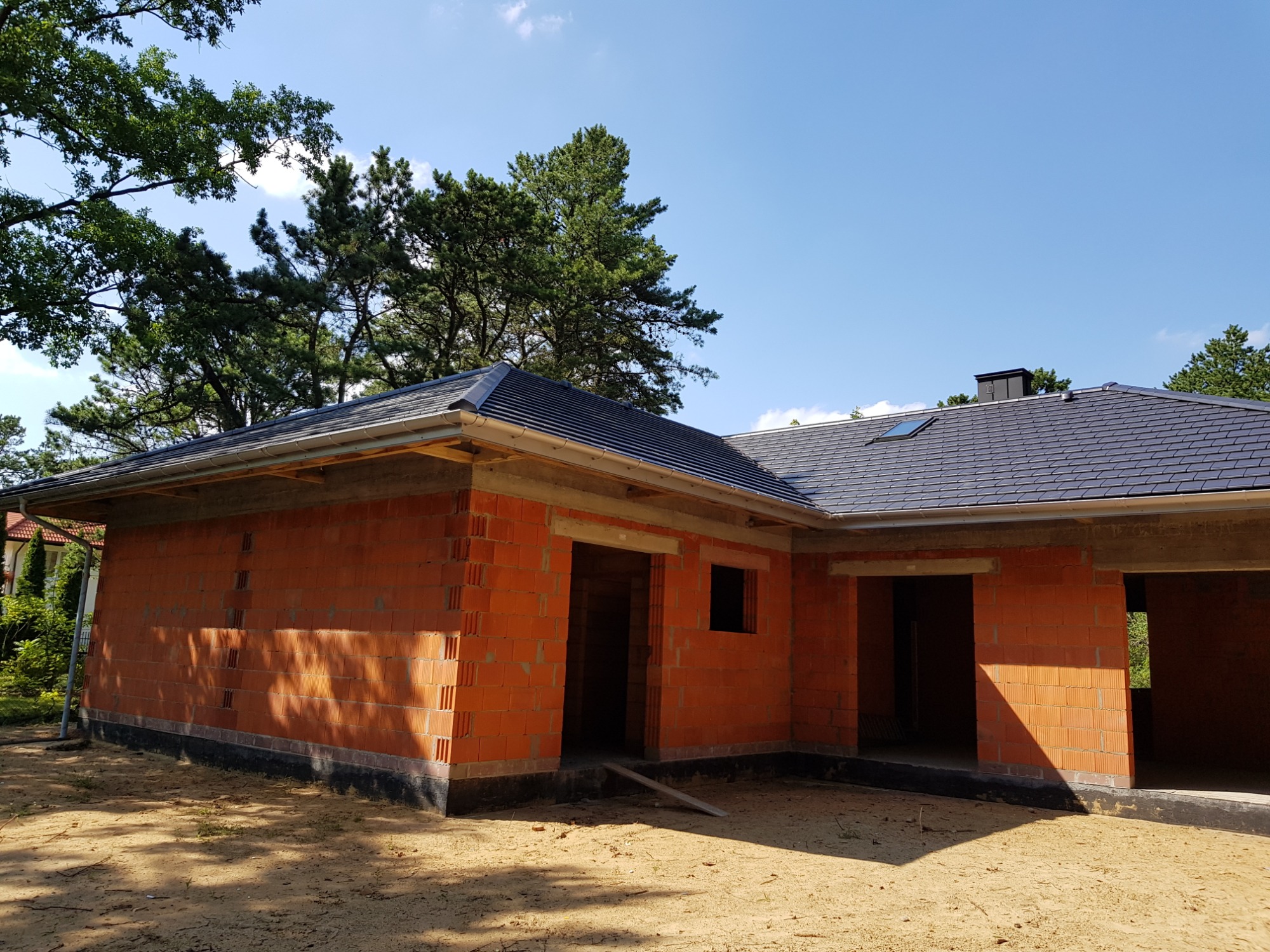 Niedokończony budynek z czerwonej cegły z zamontowanym ciemnoszarym pokryciem dachowym, widoczne rynny i komin, otoczony drzewami i piaskiem.