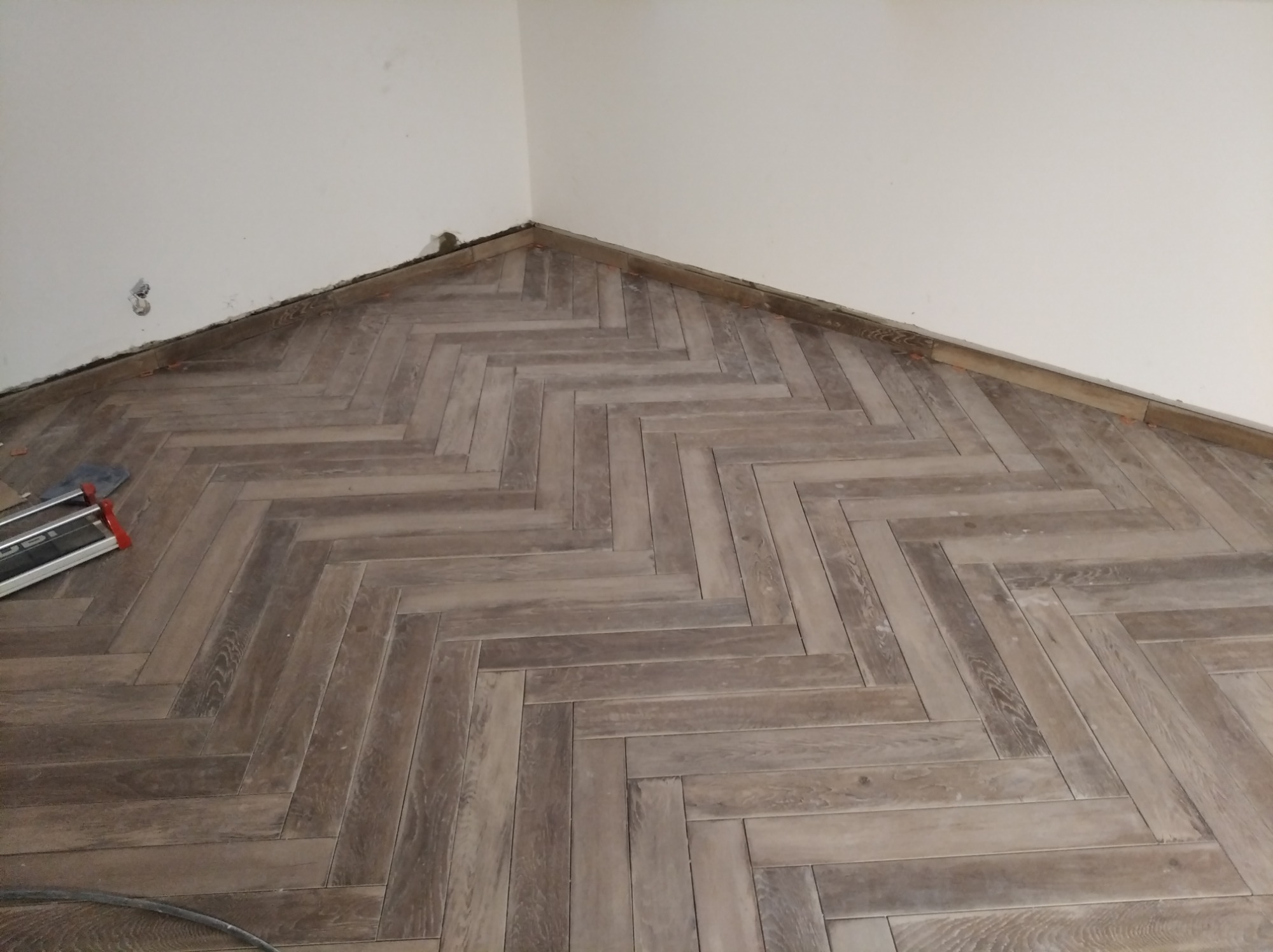 Ukończona podłoga z paneli ułożonych w jodełkę, widoczne listwy przypodłogowe w trakcie montażu, fragment narzędzia glazurniczego w lewym dolnym rogu, surowy stan ściany.