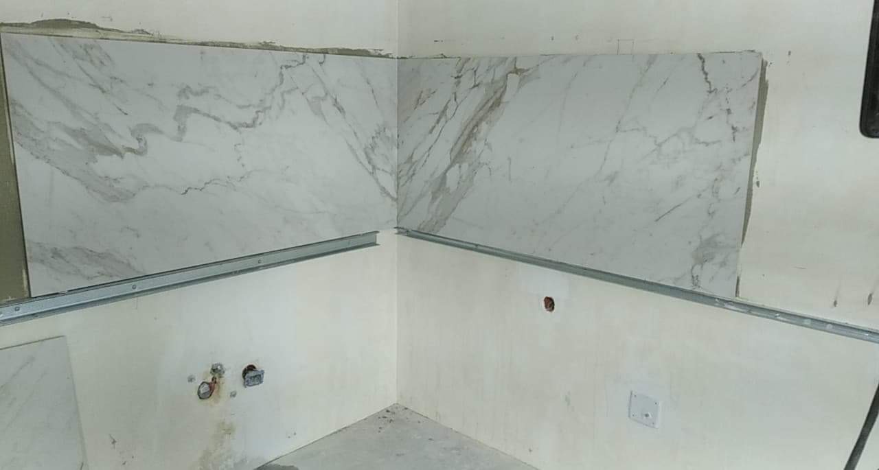 Fragment ściany w trakcie układania płytek imitujących marmur, widoczne metalowe listwy montażowe, ściana częściowo otynkowana z widocznymi puszkami elektrycznymi.