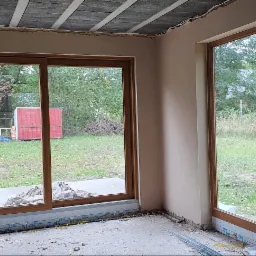 Wnętrze w trakcie prac wykończeniowych: ściany po nałożeniu wstępnej warstwy gładzi, widoczne drewniane ramy okienne, surowy betonowy sufit i elementy instalacji elektrycznej.