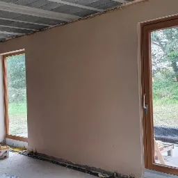 Wnętrze w trakcie tynkowania maszynowego: gładkie, beżowe ściany z widoczną maszyną do tynkowania, surowy sufit z betonowych płyt i wystającymi kablami, duże okno z drewnianą ramą i widokiem...