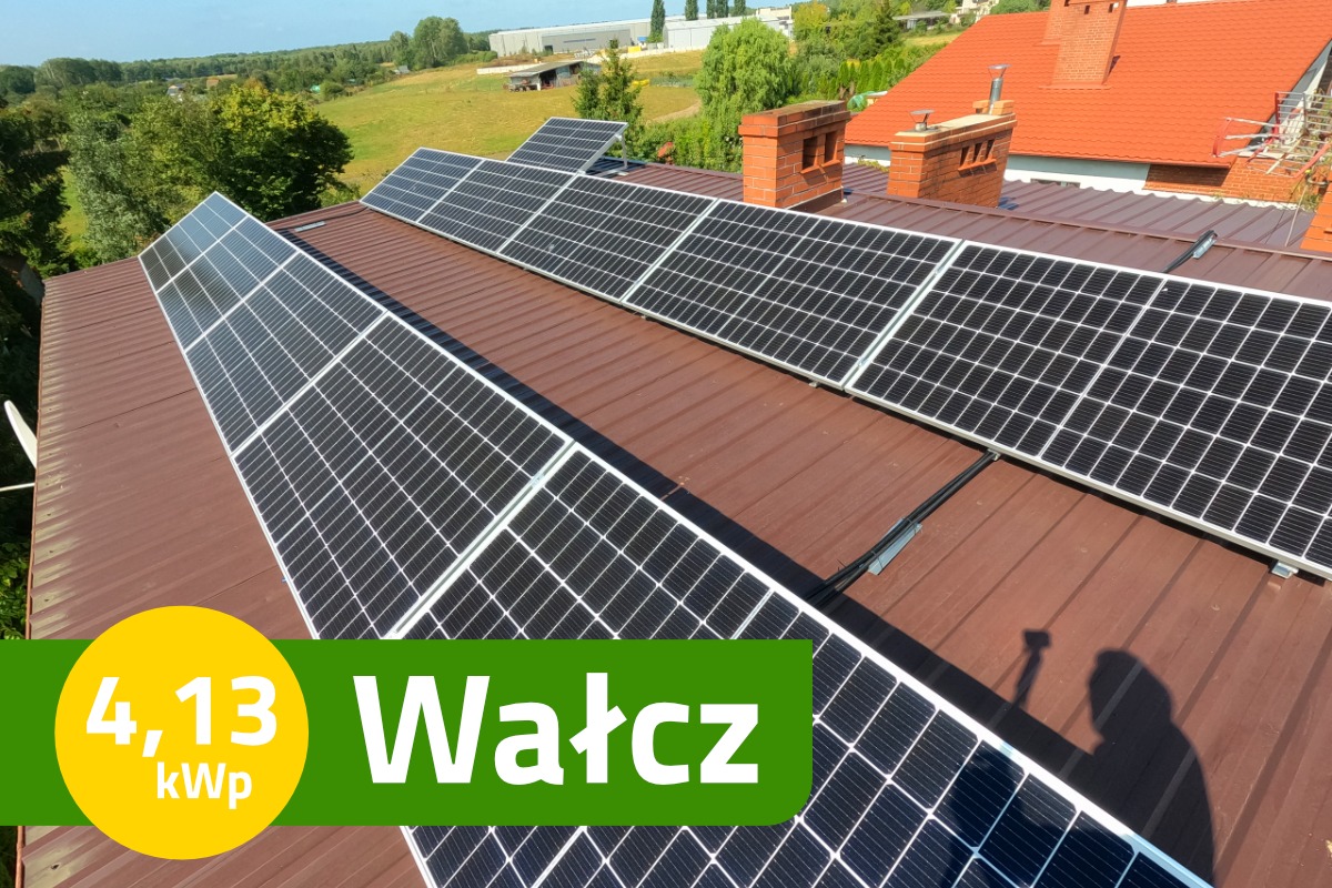 Panele słoneczne na brązowym dachu z blachy trapezowej, widok z góry, cień fotografa w prawym dolnym rogu, w tle zabudowania i zieleń, naklejka z mocą instalacji i nazwą miejscowości.