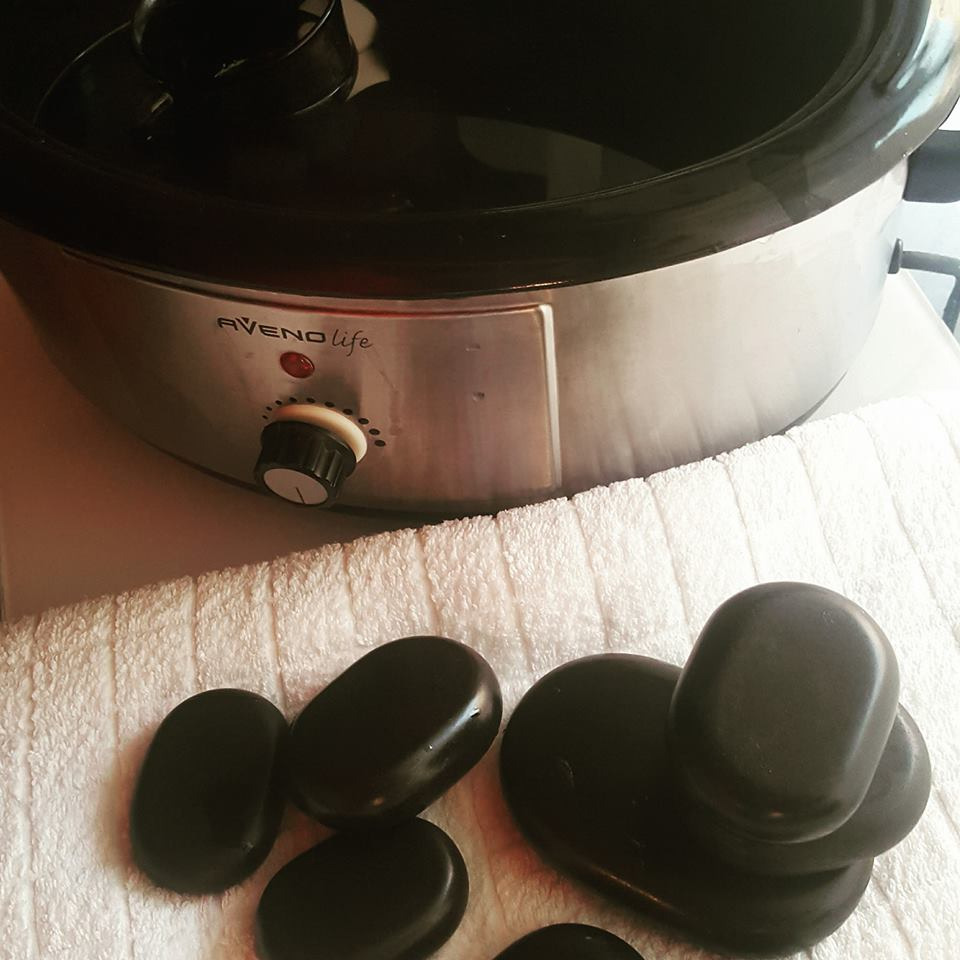 Podgrzewacz do kamieni marki Aveno Life, ustawiony na białym ręczniku obok ułożonych czarnych, gładkich kamieni do masażu.