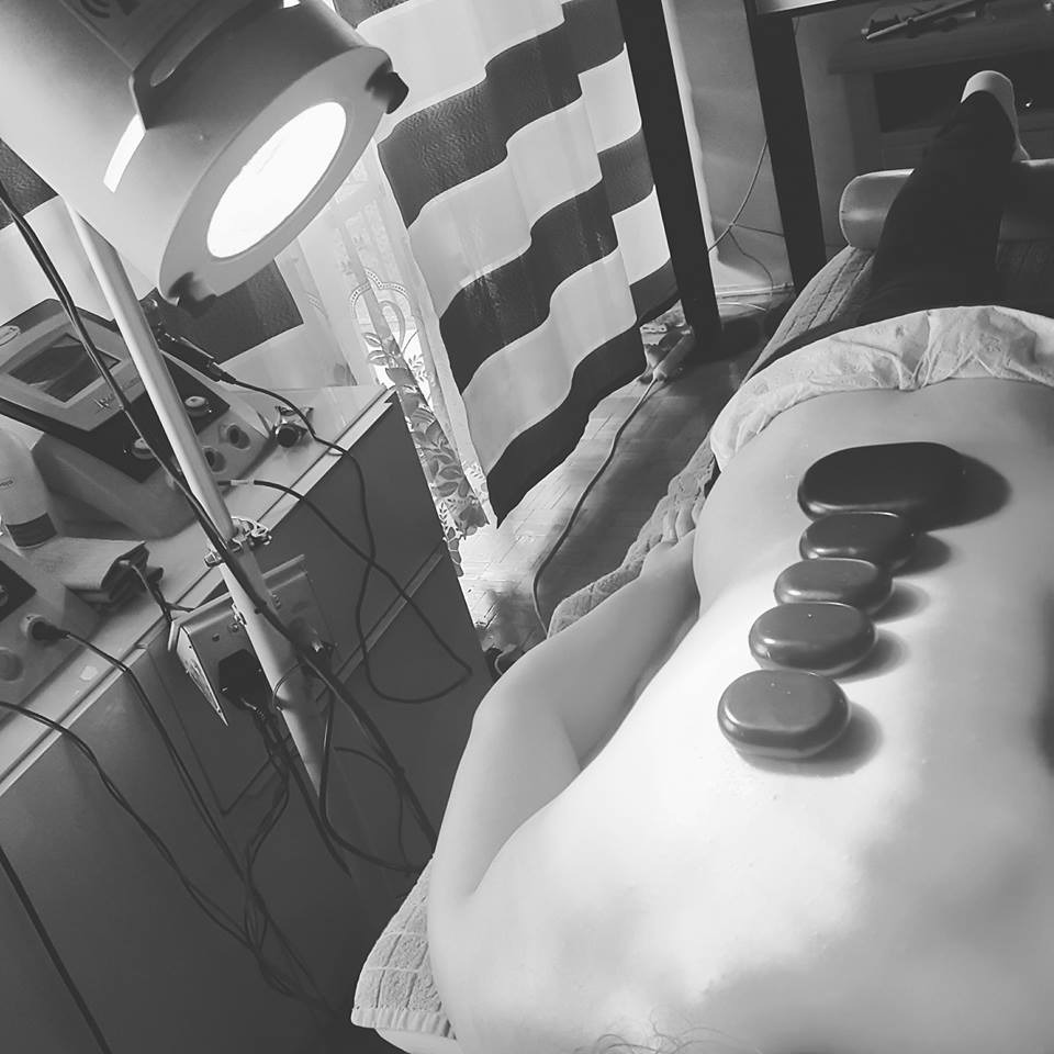 Czarno-białe zdjęcie pleców osoby leżącej na leżance z ułożonymi na kręgosłupie rozgrzanymi kamieniami bazaltowymi, obok widoczna lampa terapeutyczna i stolik z aparaturą.