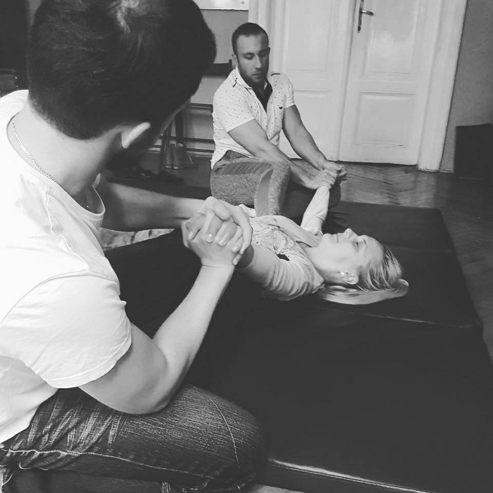 Czarno-białe zdjęcie z sesji relaksacyjnej: mężczyzna w jeansach klęczy, trzymając dłonie leżącej na macie blondynki, drugi mężczyzna siedzi w tle, przy drzwiach.