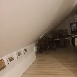 Poddasze w Szczecinie z ukośnym sufitem, jasnymi panelami podłogowymi i białymi meblami, na których stoją obrazy w ramkach. W tle widoczne rowery i regał z książkami.