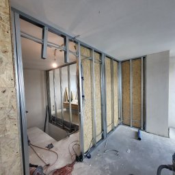 ZANE DESIGN SPÓŁKA Z OGRANICZONĄ ODPOWIEDZIALNOŚCIĄ - Wnętrze w trakcie budowy ścian działowych: metalowy szkielet, płyty OSB, widoczne przewody elektryczne, narzędzia na podłodze, surowy beton.