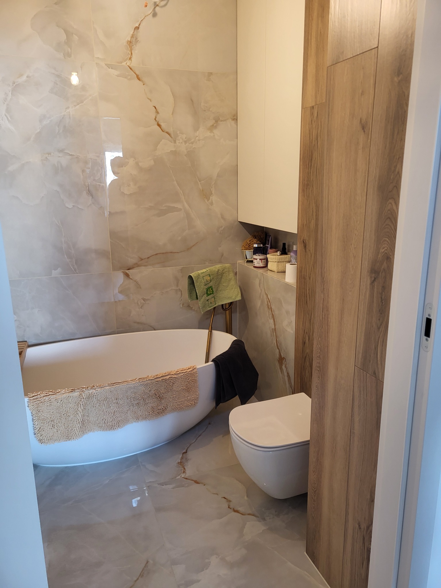 Nowoczesna łazienka z wolnostojącą wanną, umywalką wiszącą i płytkami imitującymi marmur na ścianach i podłodze, uzupełniona drewnianymi panelami.