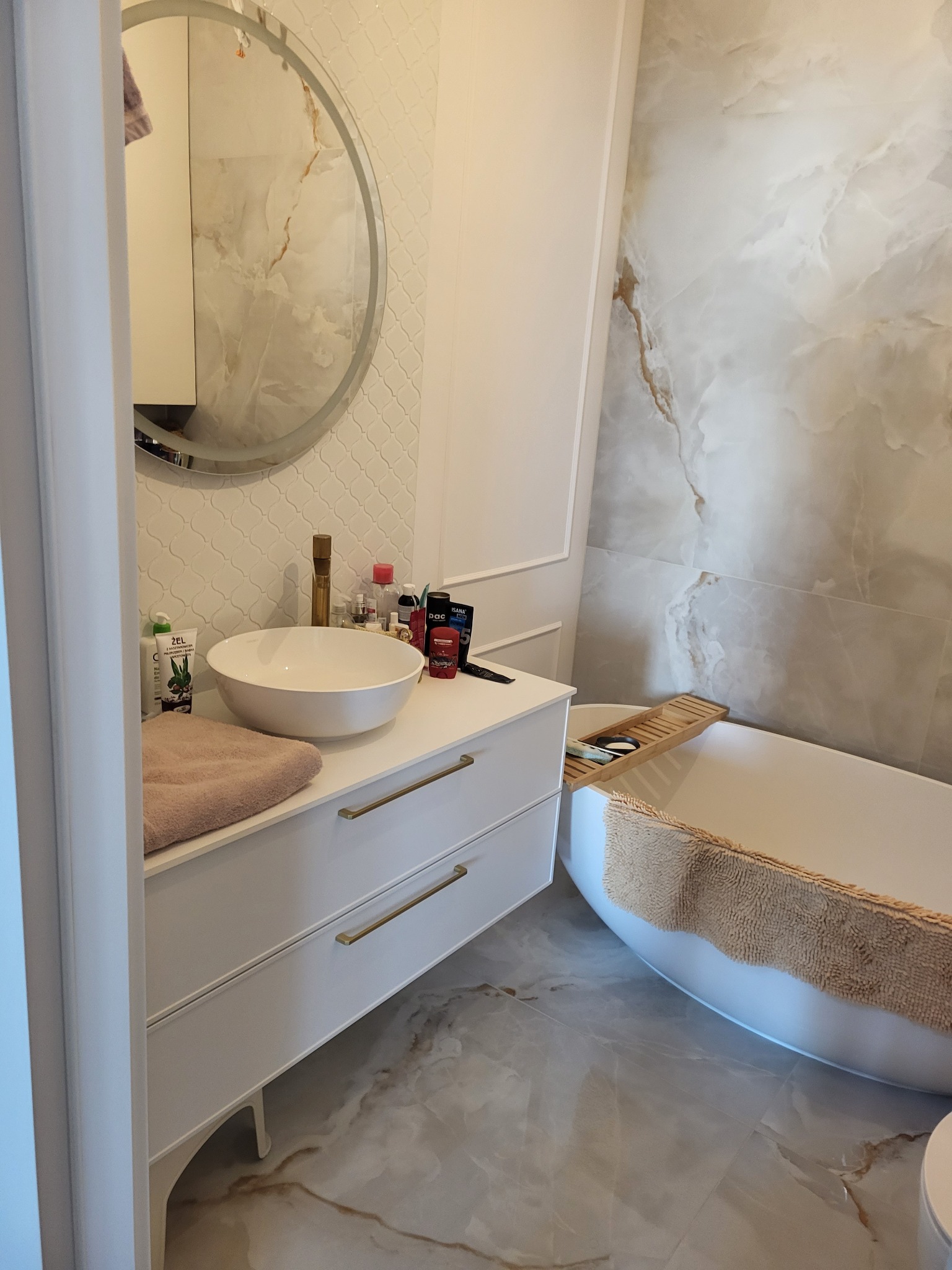 Nowoczesna łazienka z owalną umywalką nablatową, białą szafką z złotymi uchwytami, okrągłym lustrem i wolnostojącą wanną z drewnianą półką na akcesoria.