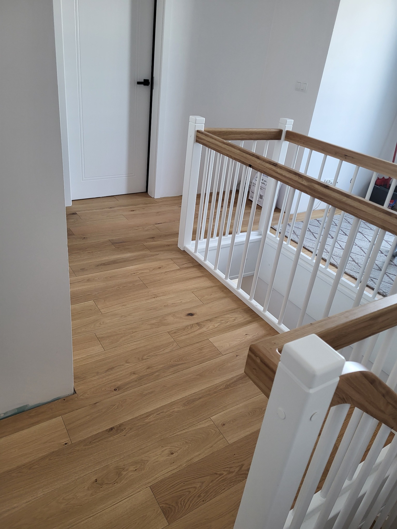 Jasna, drewniana podłoga ułożona wzdłuż korytarza z białymi drzwiami i balustradą schodów z drewnianymi poręczami.
