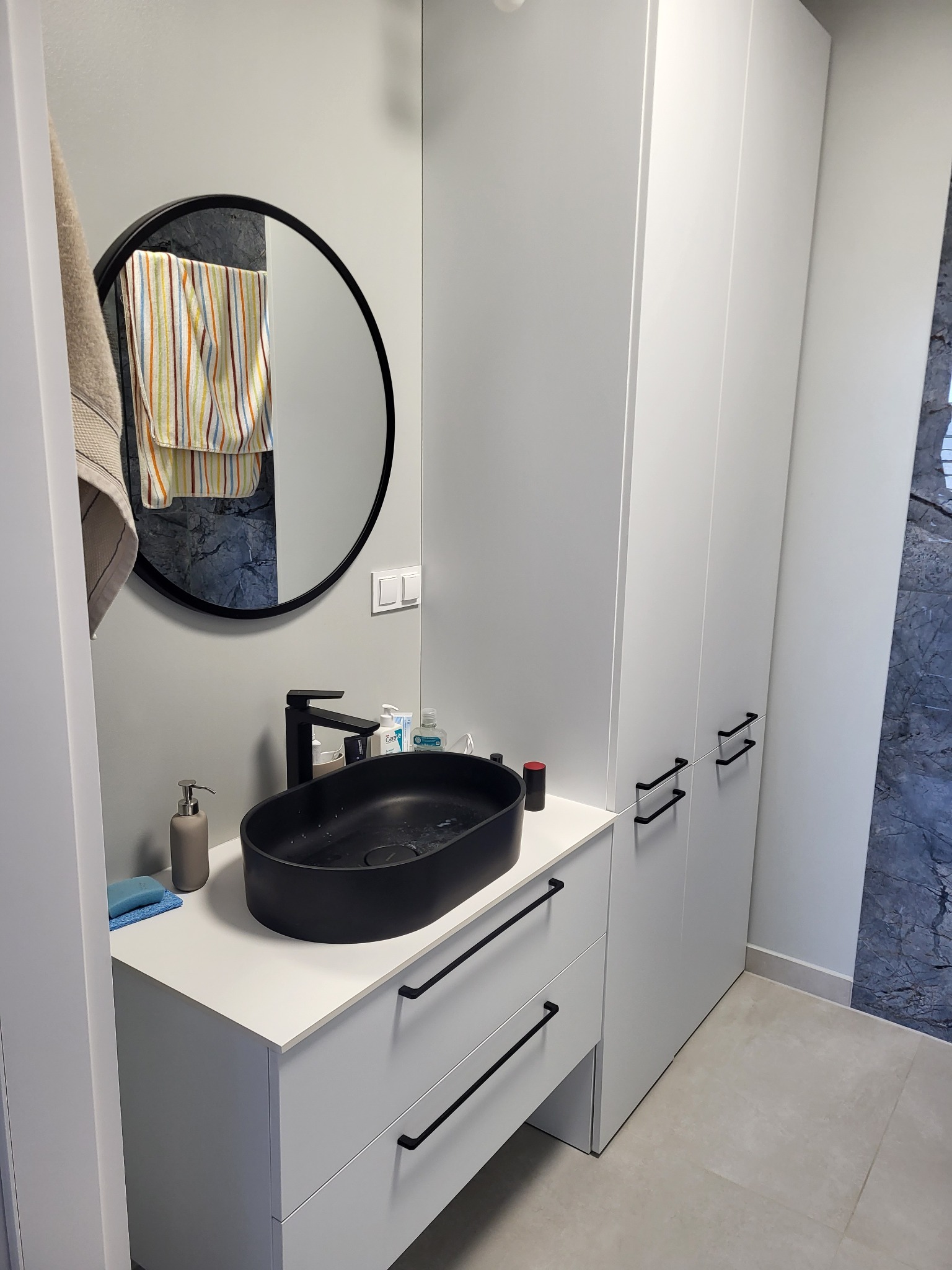 Nowoczesna łazienka w szarościach z czarną, owalną umywalką nablatową i okrągłym lustrem z czarną ramą; widoczna szafka pod umywalką z szufladami i wysoka szafa.