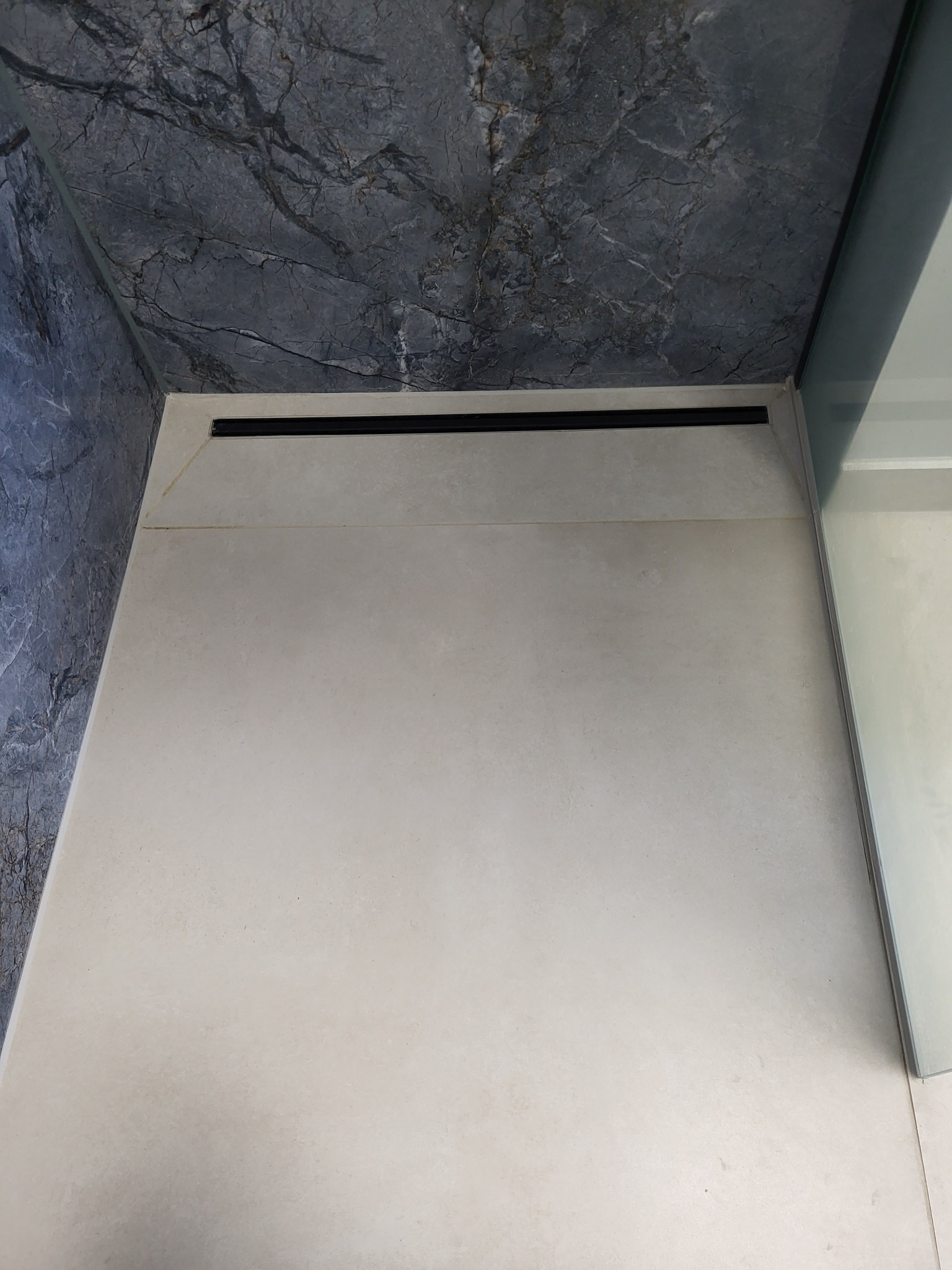 Wykończona kabina prysznicowa z odpływem liniowym, ściany wyłożone płytkami imitującymi marmur, jasne płytki podłogowe, szklana ścianka kabiny.