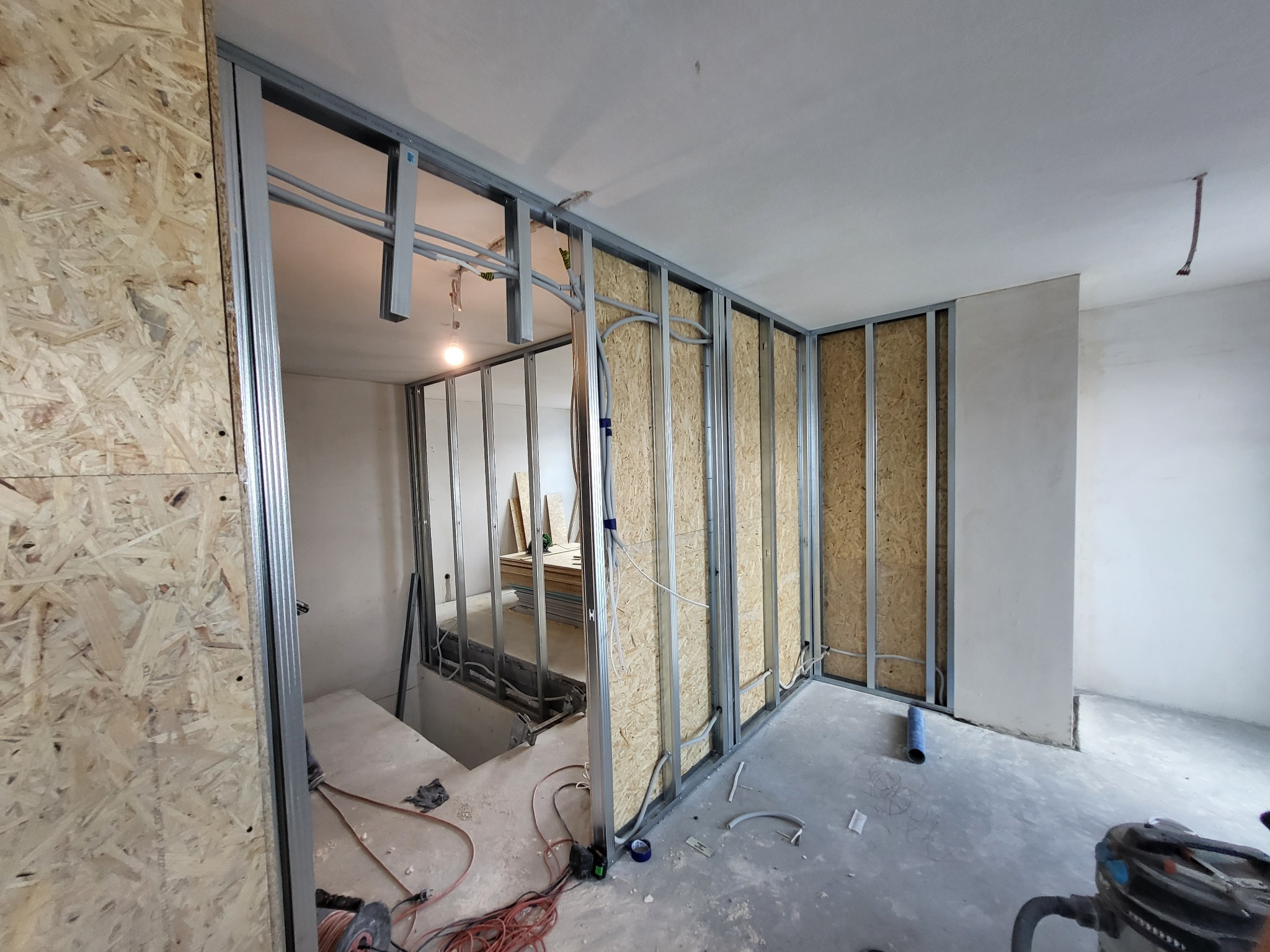 Wnętrze w trakcie budowy ścian działowych: metalowy szkielet, płyty OSB, widoczne przewody elektryczne, narzędzia na podłodze, surowy beton.