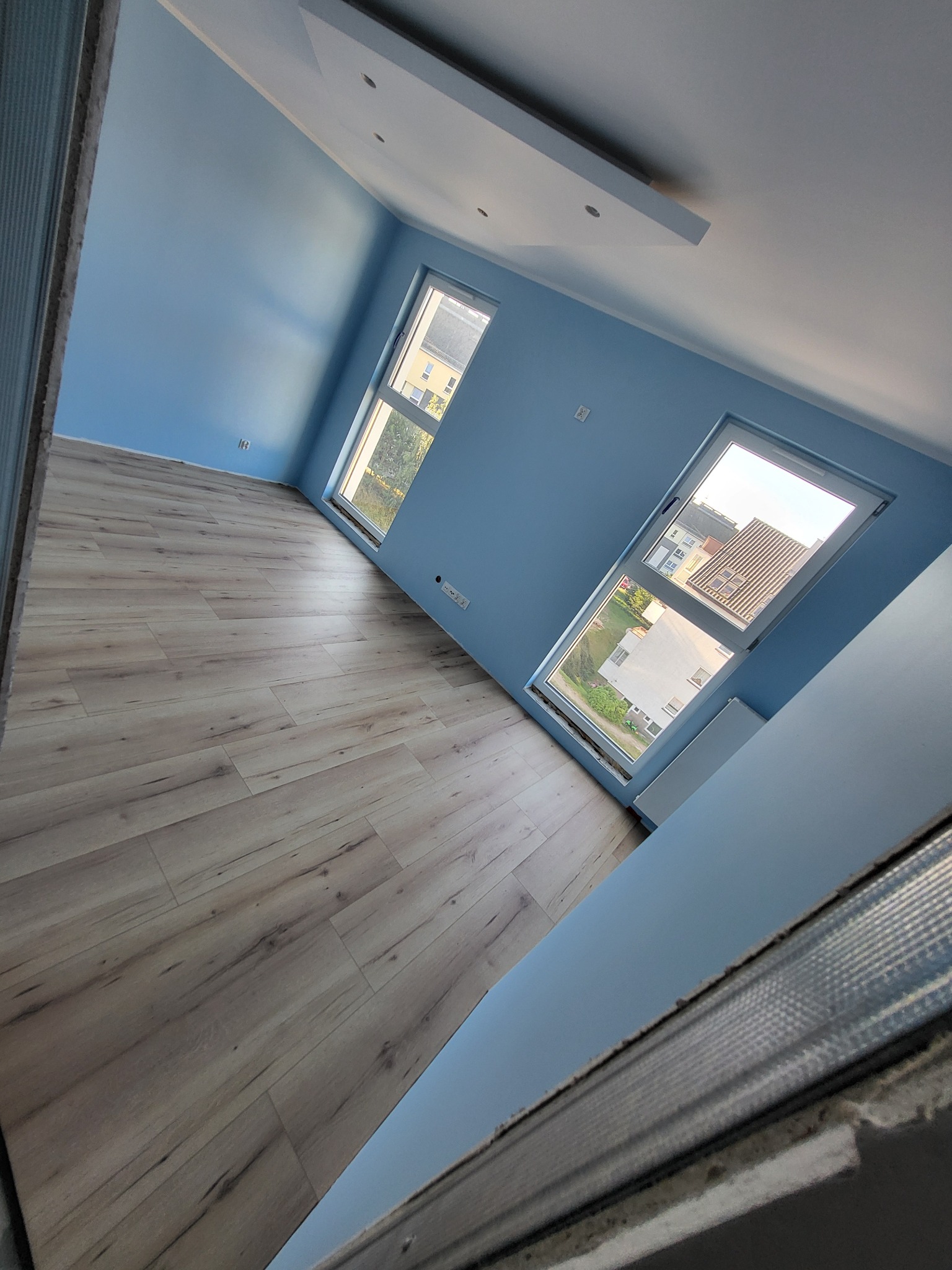 Pomalowane na niebiesko wnętrze pokoju z ułożonymi panelami podłogowymi w odcieniach drewna, widoczne dwa okna z grzejnikami pod nimi oraz fragment sufitu z oświetleniem.