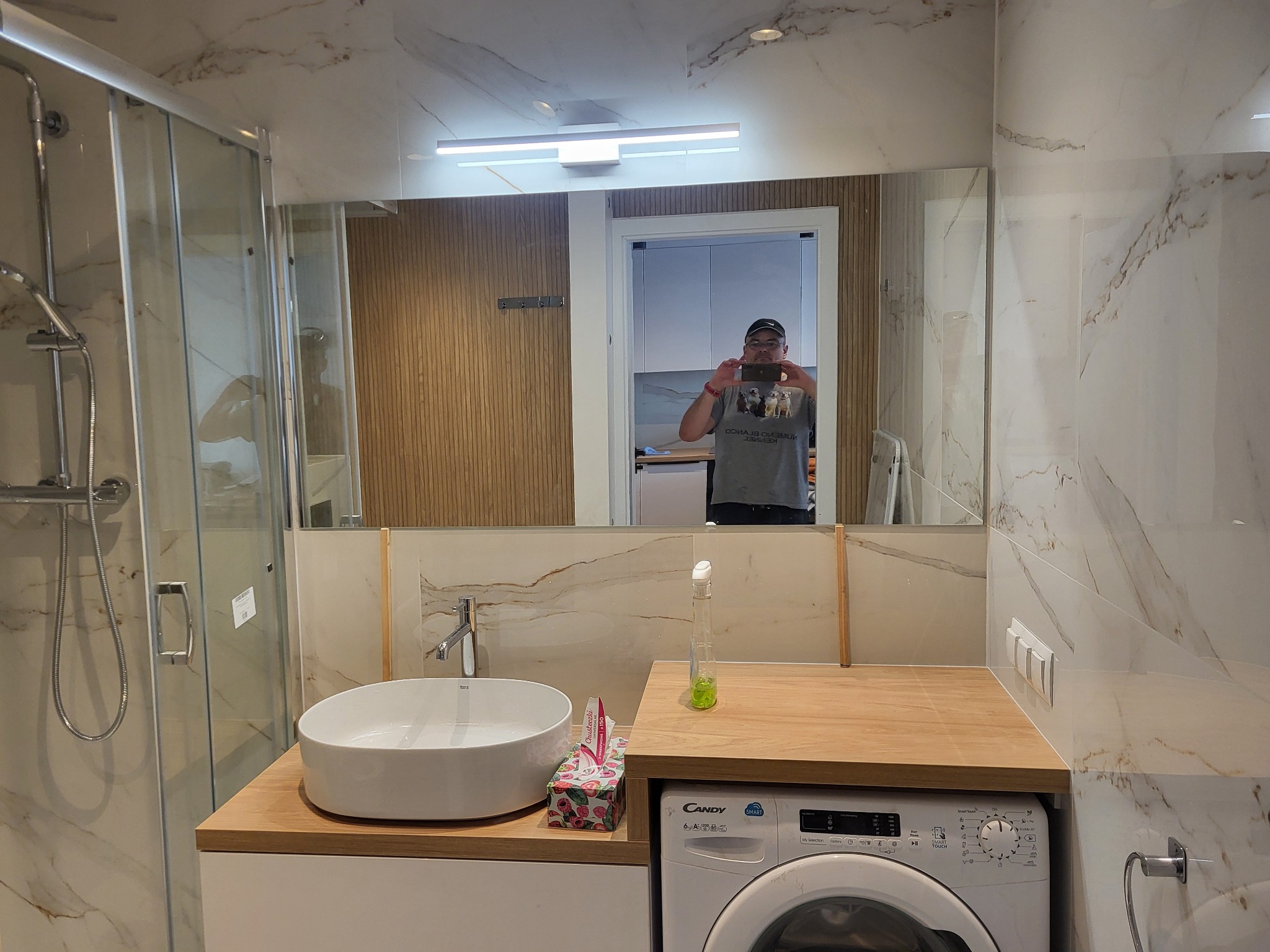 Nowoczesna łazienka z marmurowymi płytkami, kabiną prysznicową, umywalką nablatową i pralką pod blatem, sfotografowana z odbiciem osoby robiącej zdjęcie w lustrze.