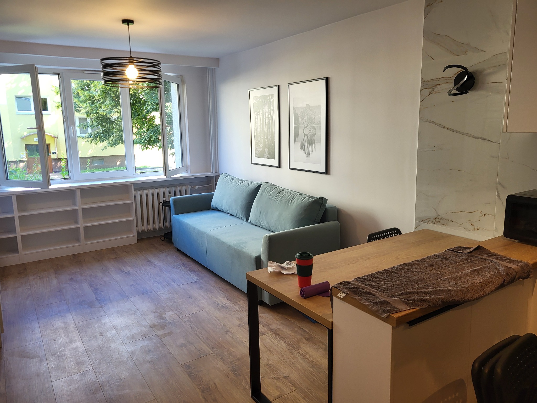 Jasne wnętrze pokoju z błękitną sofą, drewnianą podłogą i stołem, widokiem z okna na zieleń i minimalistycznymi dekoracjami ściennymi.