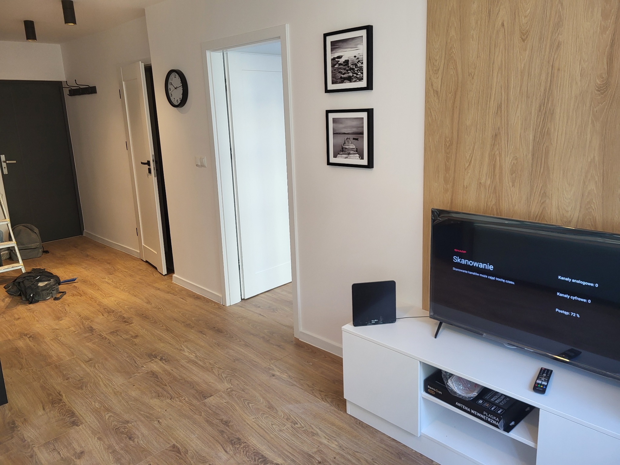 Jasne panele podłogowe w nowoczesnym mieszkaniu, widoczny fragment ściany z okładziną drewnopodobną, biała szafka RTV z telewizorem i dekoracjami ściennymi, otwarte białe drzwi w głębi.