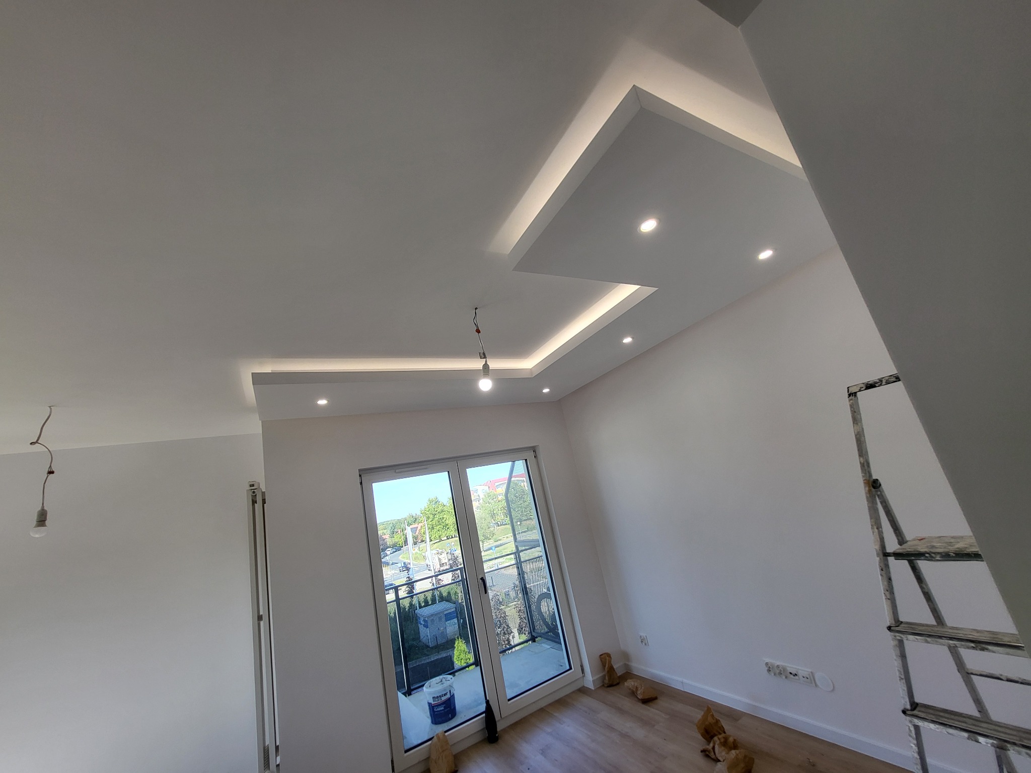 Nowoczesne wykończenie wnętrza: białe ściany, podwieszany sufit z oświetleniem LED, okno balkonowe z widokiem na zewnątrz, drabina malarska i niedokończone podłączenie lampy.