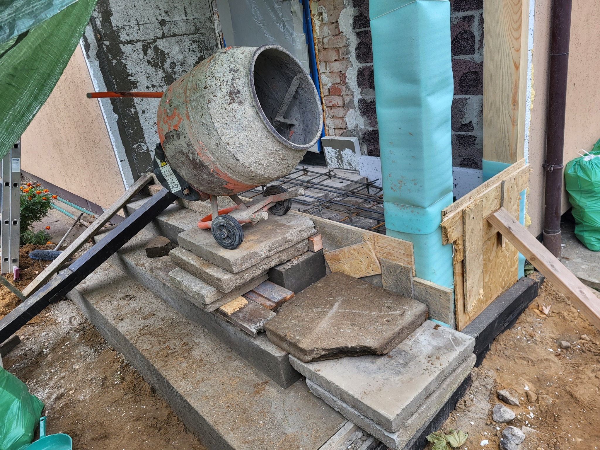 Betoniarka ustawiona na schodach przed wejściem do budynku w trakcie prac remontowych, widoczne materiały budowlane i stalowe zbrojenie.