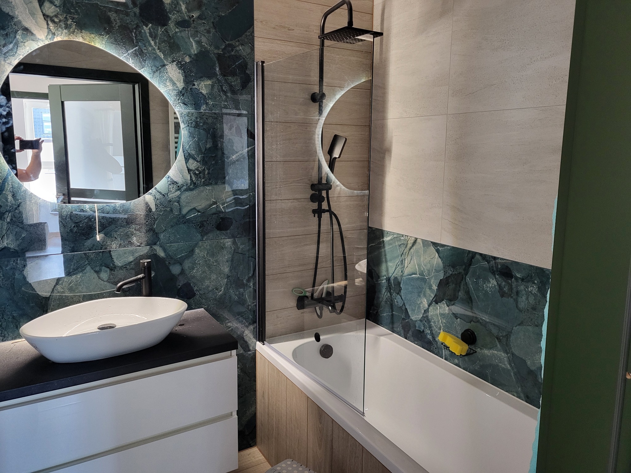 Nowoczesna łazienka z owalnym lustrem podświetlanym LED, umywalką nablatową, czarną armaturą i wanną za szklaną ścianką; płytki imitujące zielony marmur na ścianach.