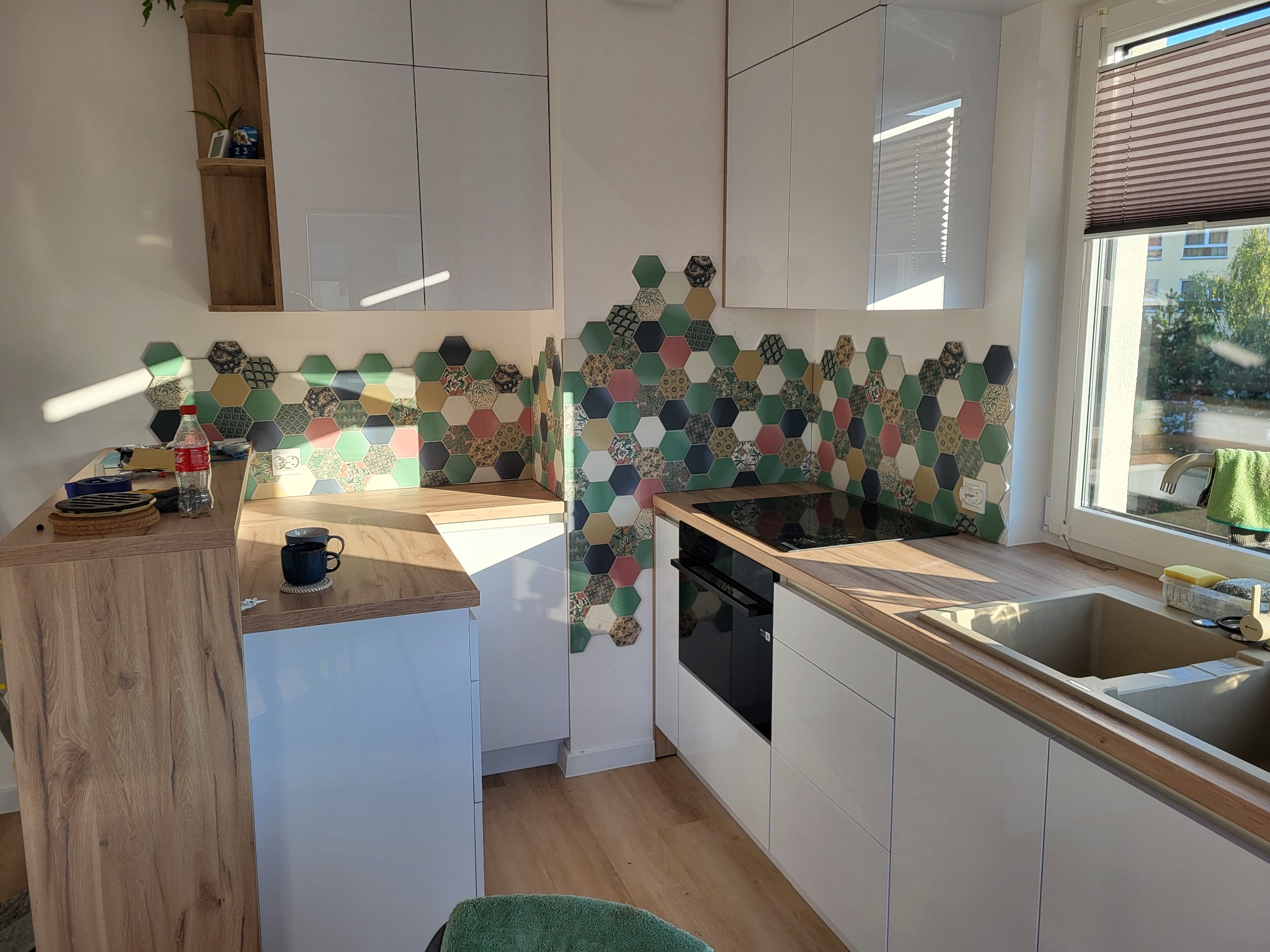 Jasna kuchnia z białymi szafkami, drewnianymi blatami i heksagonalnymi płytkami dekoracyjnymi nad blatem, widok na okno z żaluzjami.