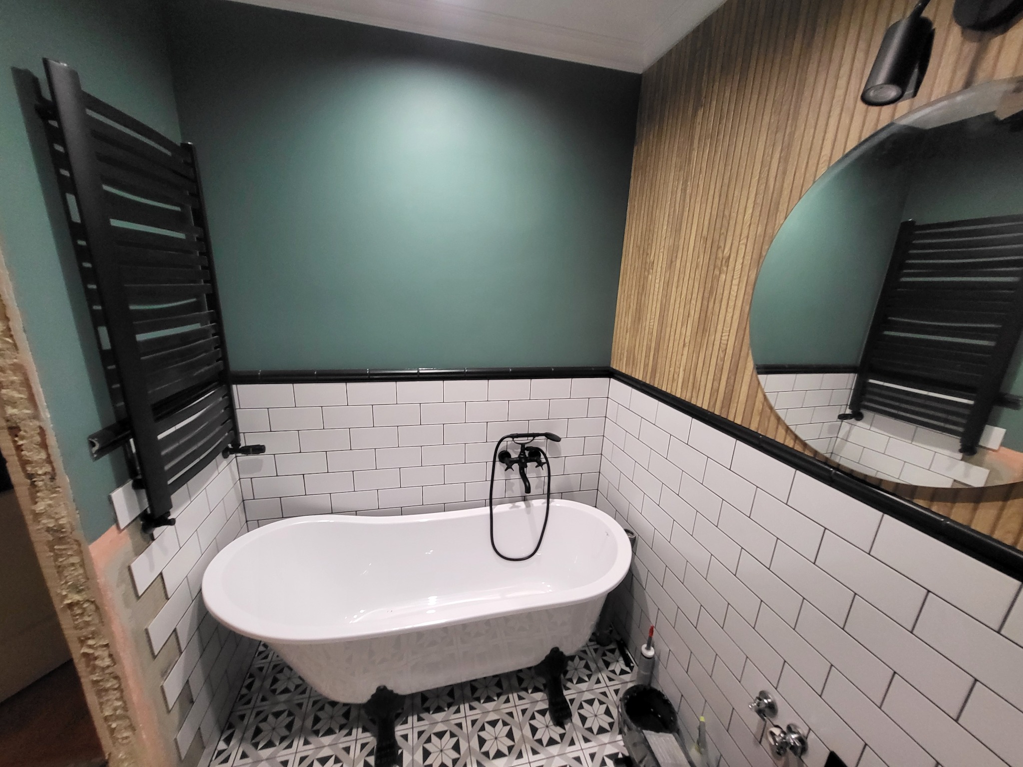 Biała wanna wolnostojąca na czarnych nóżkach w łazience z białą cegiełką, zieloną ścianą i drewnianą okładziną, widoczny czarny grzejnik drabinkowy i wzorzysta podłoga.