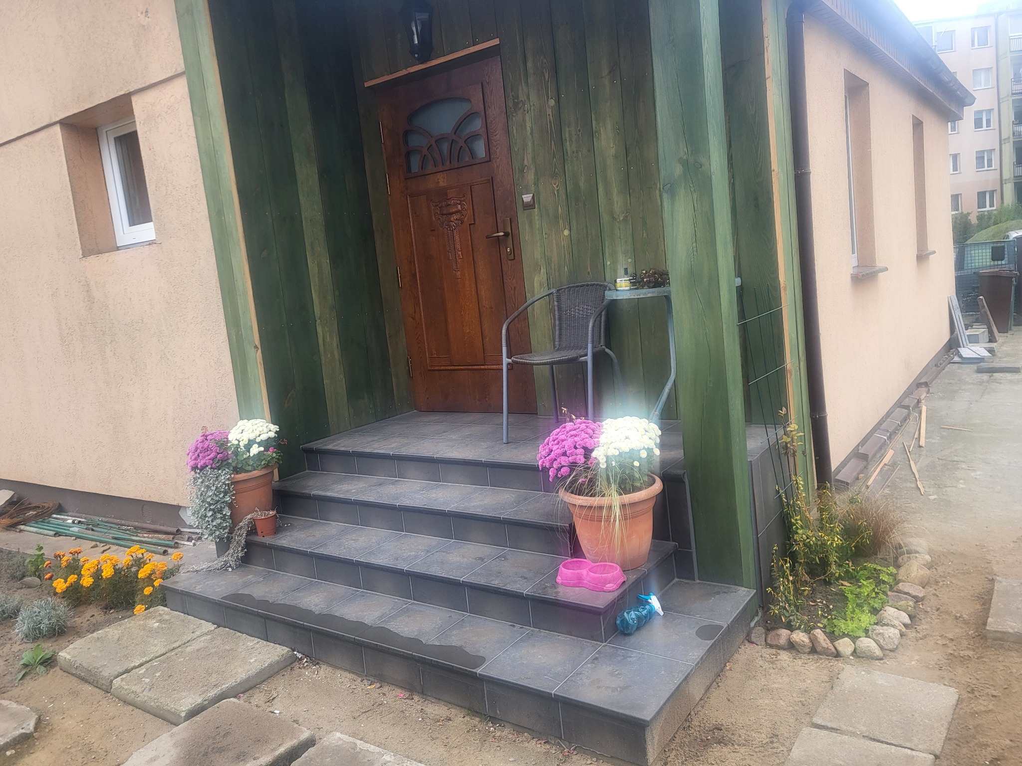 Wejście do domu z drewnianymi drzwiami i schodami wyłożonymi ciemnymi płytkami, ozdobione kwiatami w donicach i elementami dekoracyjnymi.