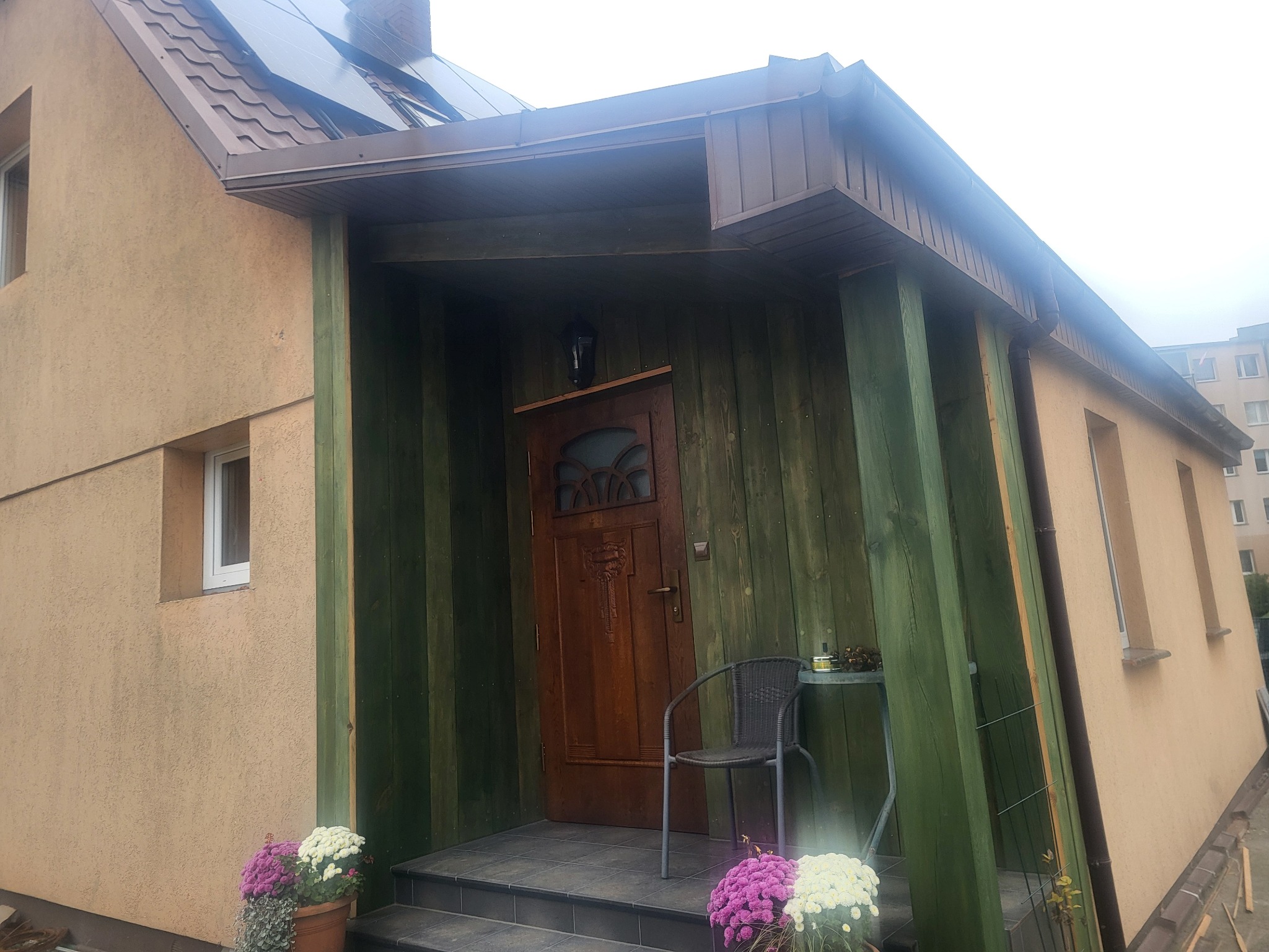 Wejście do domu z drewnianą werandą pomalowaną na zielono, brązowe drzwi z dekoracyjnym przeszkleniem, kwiaty w donicach i krzesło z małym stolikiem przed wejściem.