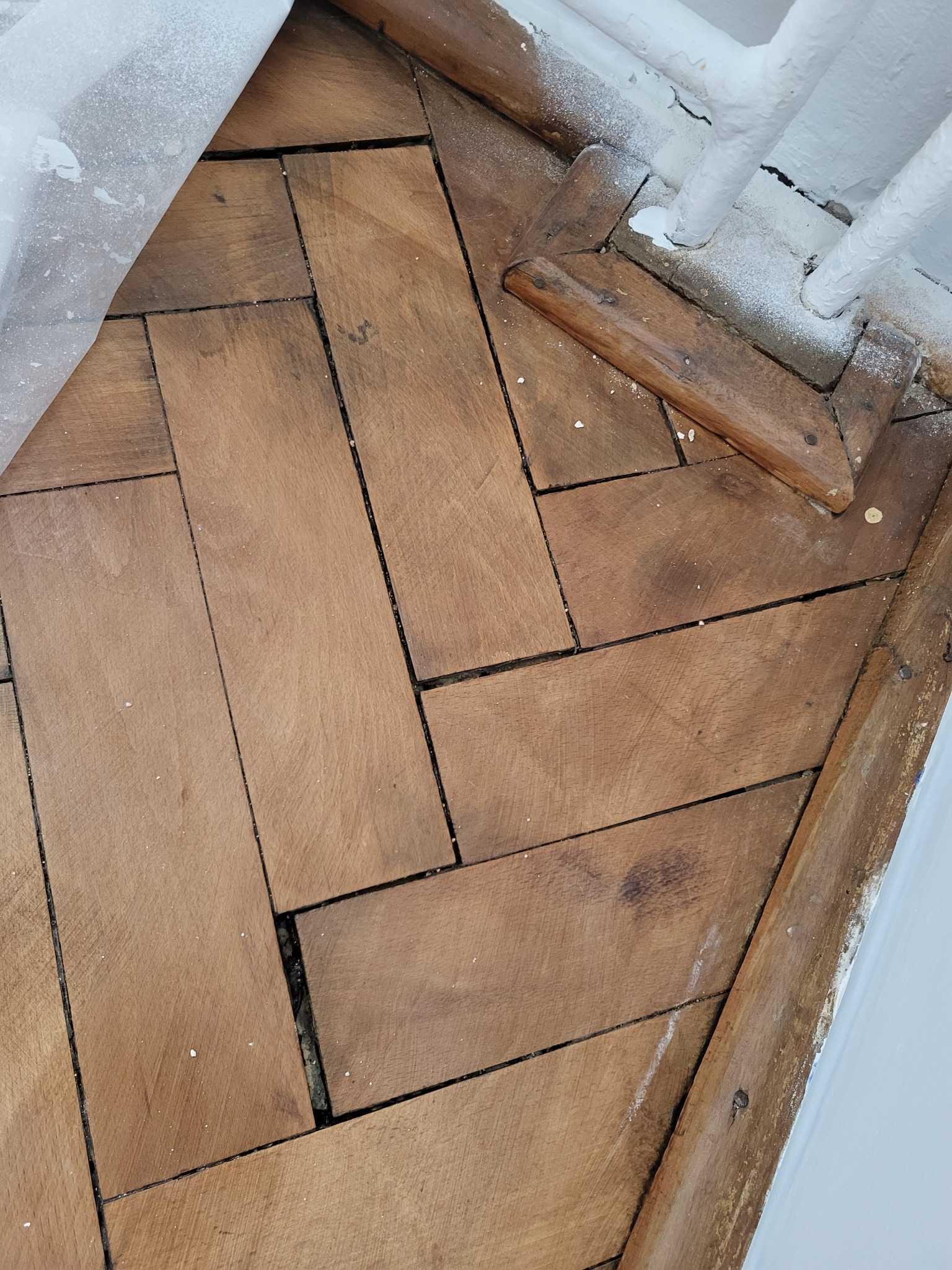 Ułożona w jodełkę, stara drewniana podłoga z widocznymi śladami użytkowania, pyłem i zabrudzeniami, fragmentem folii malarskiej i pomalowaną na biało barierką.