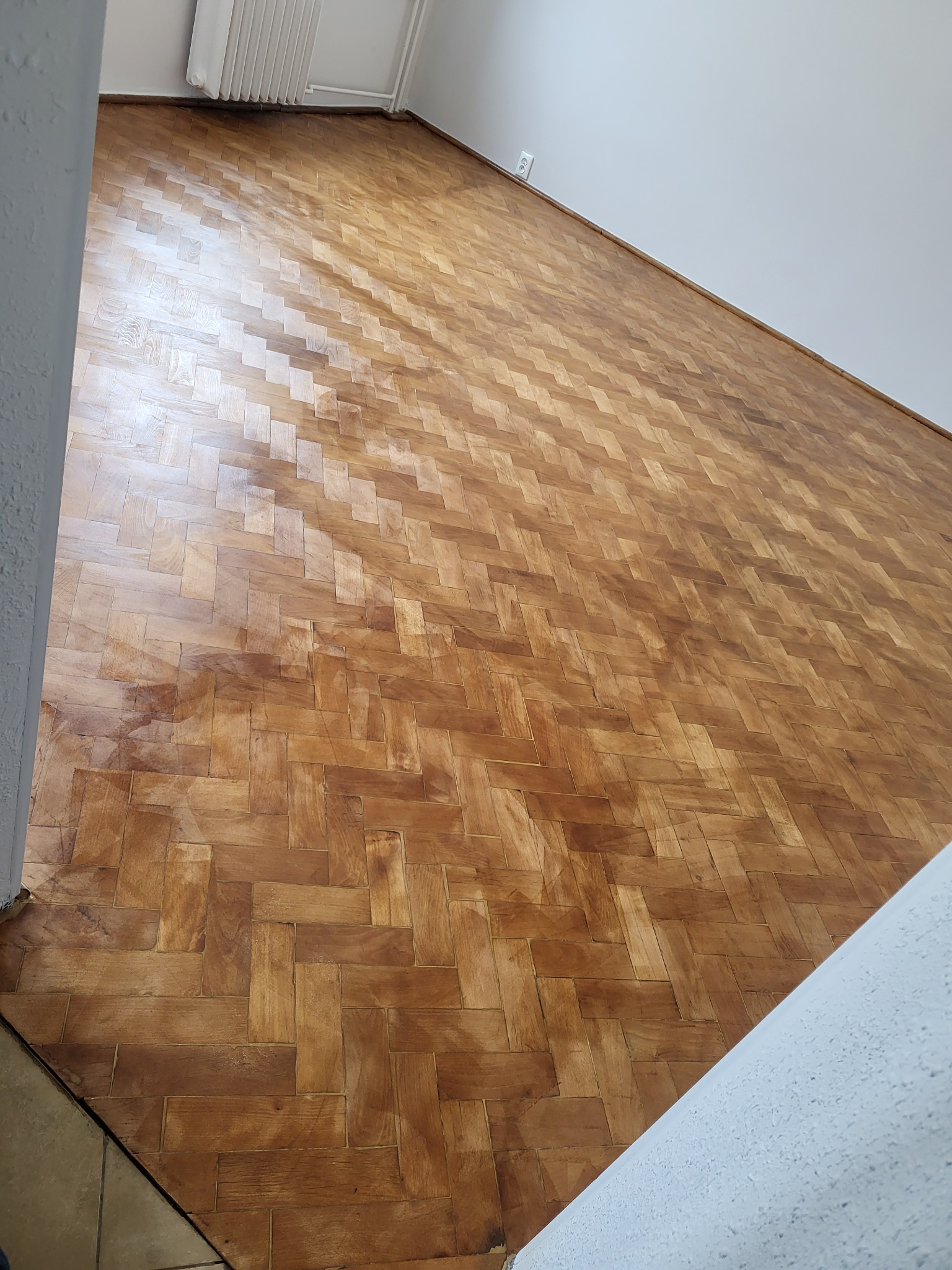 Ułożona w jodełkę, lakierowana parkietowa podłoga z widocznym fragmentem ściany i grzejnika w tle.