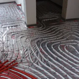 Rozłożone rury ogrzewania podłogowego na folii izolacyjnej z rastrem, widoczne czerwone rury doprowadzające i czarne uchwyty mocujące, w tle surowy stan wykończenia wnętrza.